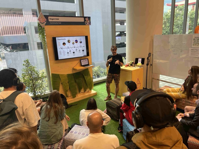 People seated on a green carpet and cushions listening to a speaker beside a screen inside a conference hall, the Regen Hub in Bangkok